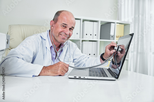 Smiling middle-aged general practitioner listening laptop screen with stethoscope