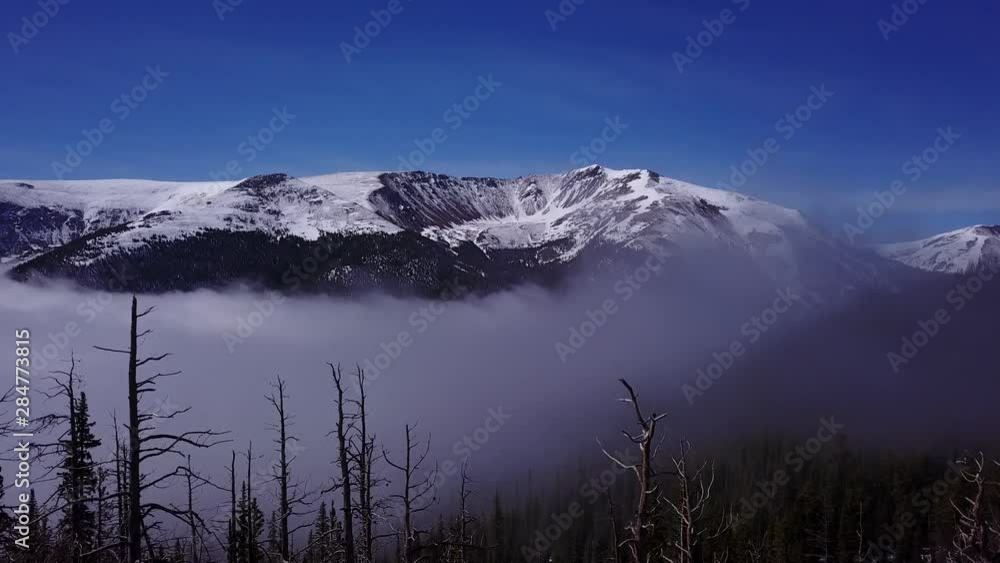 Flying above the clouds in the Rocky Mountains. Aerial footage flying over trees to reveal a foggy valley beneath snow capped mountains. Flying above the clouds in the Rocky Mountains. Aerial footage flying over trees to reveal a foggy valley beneath snow capped mountains.