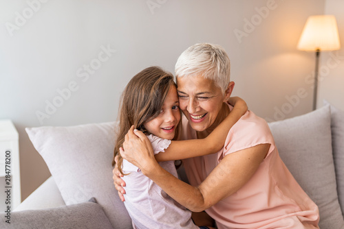 Cute little girl having fun playing with smiling grandmother embracing lying on couch together, happy granny and preschool grandchild relax on sofa, grandma granddaughter cuddling laughing at home