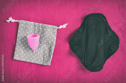 Reusable eco-friendly feminine hygiene period products - menstrual cup and black organic cotton menstruation pad - cloth menstrual pads to wash. Top view on red towel background.