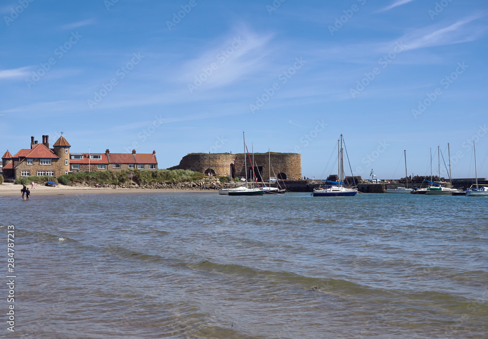 Looking North towards the Harbour at Beadnell Bay, with people Paddling in the Sea, and Yachts anchored next to the Breakwater, Northumberland, England.