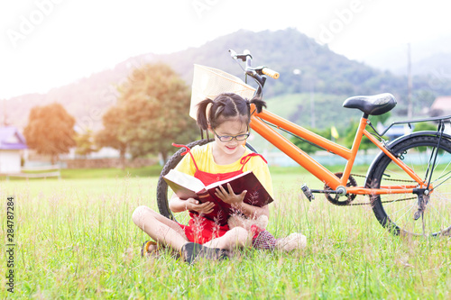 Cute asian student girl with glasses sitting on the grass reading a book in garden at summertime