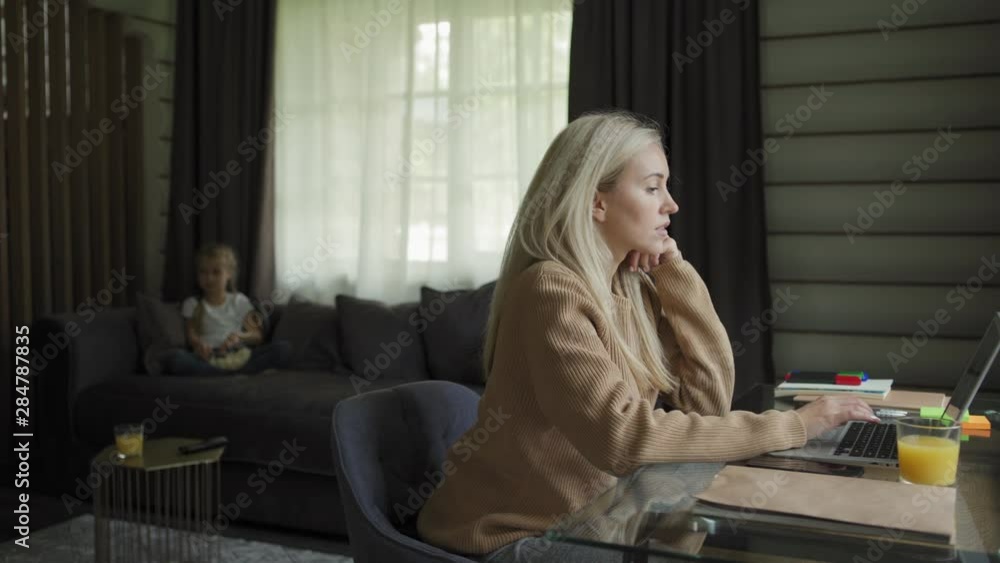 Tracking right shot of blonde entrepreneur woman working on laptop computer sitting at desk at home, her little daughter watching tv and eating popcorn in background Tracking right shot of blonde entrepreneur woman working on laptop computer sitting at desk at home, her little daughter watching tv and eating popcorn in background