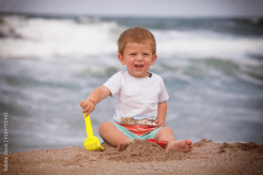 A cute little boy is sitting on the sea sand. Happy little boy on the beach. Summer. Recreation. Tourism. A cute little boy is sitting on the sea sand. Happy little boy on the beach. Summer. Recreation. Tourism.