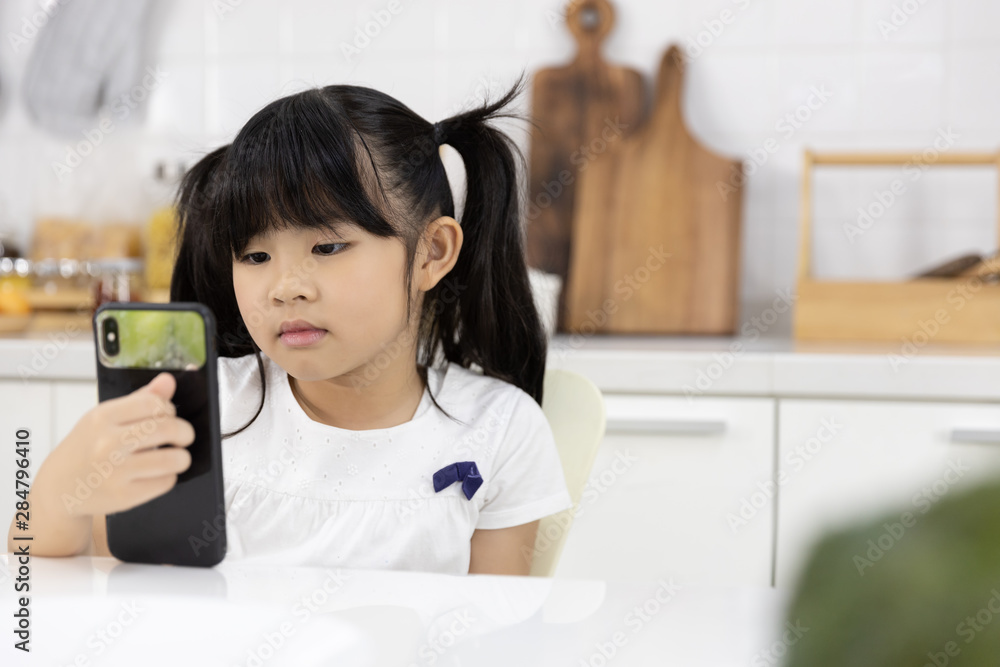 Happy Asian Little girl enjoy smart phone while waiting for the mother preparing breakfast in the kitchen at home. Technology and everyday life Happy Asian Little girl enjoy smart phone while waiting for the mother preparing breakfast in the kitchen at home. Technology and everyday life
