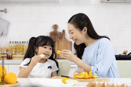 Happy Asian family Little girl eating Orange fruit and Mother are preparing the vegetables and fruit in the kitchen at home. Healthy food concept