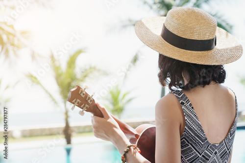 Happy woman relaxing and playing ukulele sitting on edge of swimming pool enjoy life on tropical beach sea in summer with blue sky.