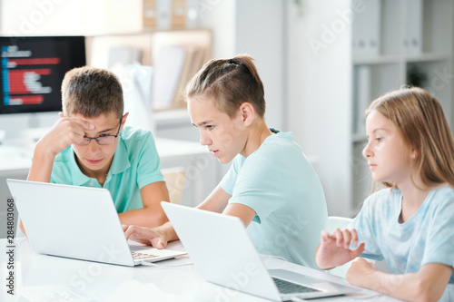 Cute schoolchildren sitting by desk in classroom while browsing in the net