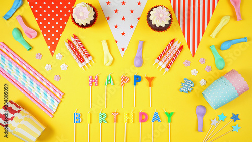 Colorful birthday party flat lay with party food, balloons, decorations and gifts on yellow background, with candles spelling Happy Birthday.