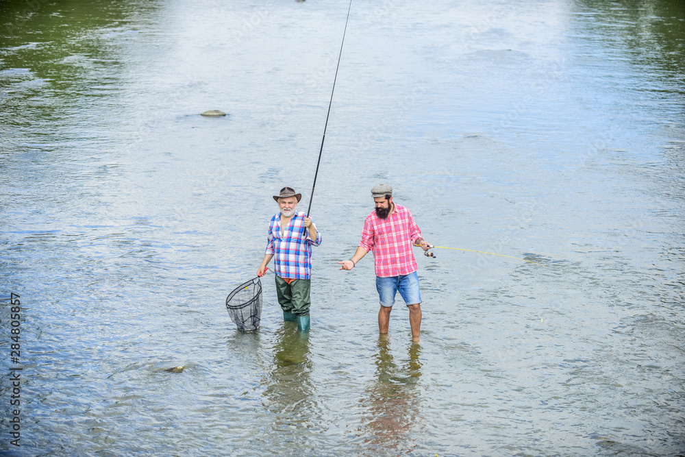 Men stand in water. Fishing is much more than fish. Happy fisherman with fishing rod and net. Hobby and sport activity. Male friendship. Father and son fishing. Summer weekend. Fishing together Men stand in water. Fishing is much more than fish. Happy fisherman with fishing rod and net. Hobby and sport activity. Male friendship. Father and son fishing. Summer weekend. Fishing together