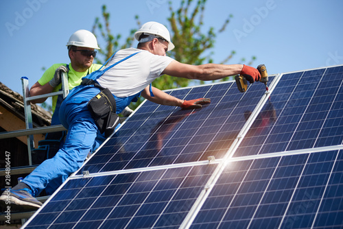 Two workers technicians installing heavy solar photo voltaic panels to high steel platform. Exterior solar system installation, alternative renewable green energy generation concept.