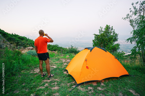 Man is drinking from cup near an orange tent standing on hill