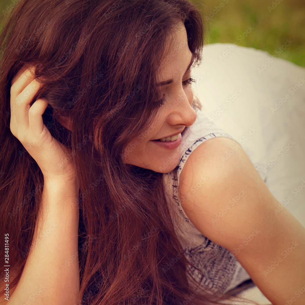Obraz premium Beautiful smiling young woman with long hair lying on the grass on summer green background. Closeup outdoor toned color bright portrait.