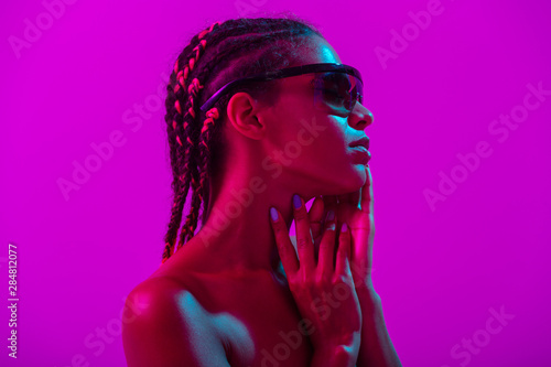 Attractive beautiful young african naked woman posing isolated over purple neon lights wall background wearing sunglasses.