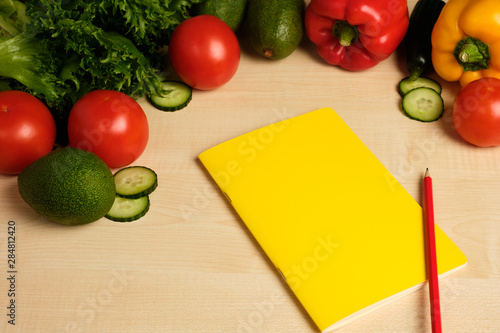 Red and green vegetables are lying on a light wooden table over a yellow notepad with a red pencil.
