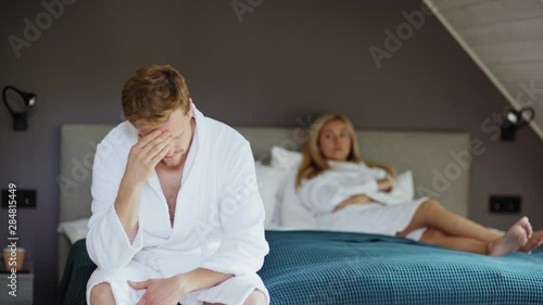 Rack focus of upset woman in white bathrobe lying on bed looking displeased. Depressed husband or lover rubbing forehead in foreground and looking at camera