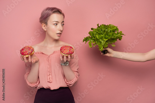 A girl with short hair in a blouse holds donuts in her hands, and looks at the salad in outstretched hand, on a pink background.