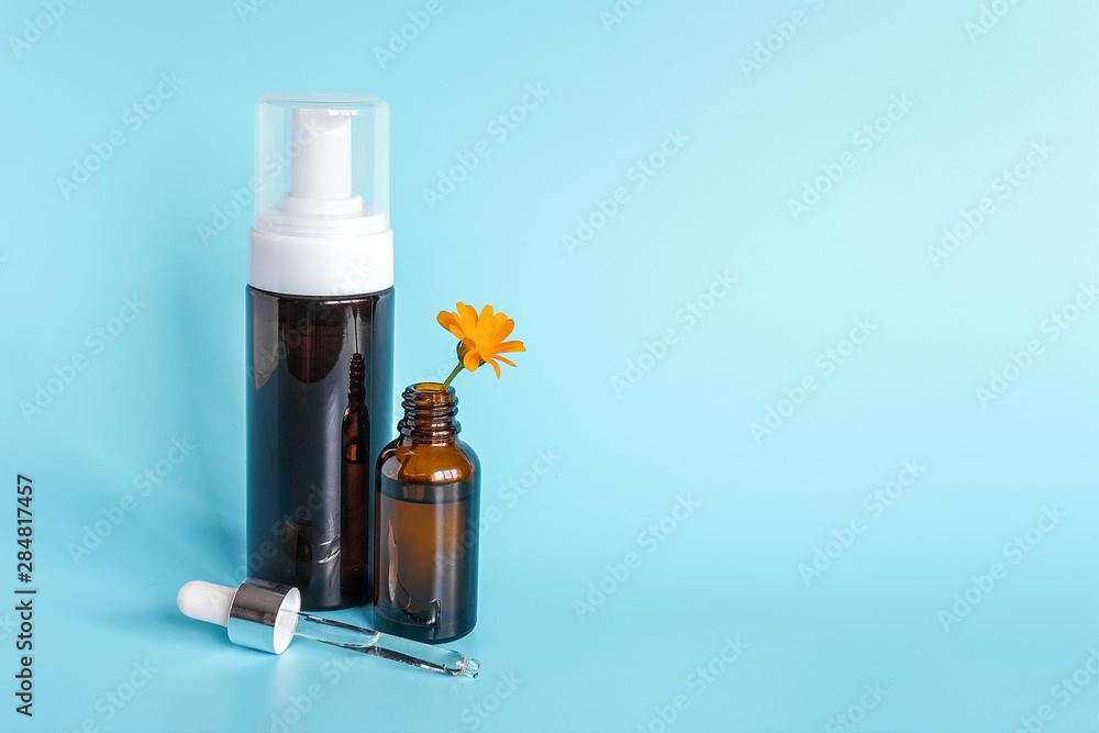 Essential oil in open brown dropper bottle with lying glass pipette, big bottle with white dispenser and orange flower calendula on blue background. Concept natural organic beauty cosmetics product Essential oil in open brown dropper bottle with lying glass pipette, big bottle with white dispenser and orange flower calendula on blue background. Concept natural organic beauty cosmetics product