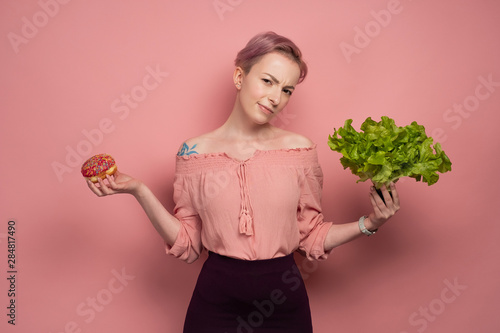 A girl with short pink hair in a blouse holds a donut and salad in her hands, looks mockingly at the camera, on a pink background photo