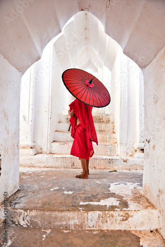 novice buddhist monks with red traditional robes holding red umbrellas walking in a white buddhist temple in myanmar
