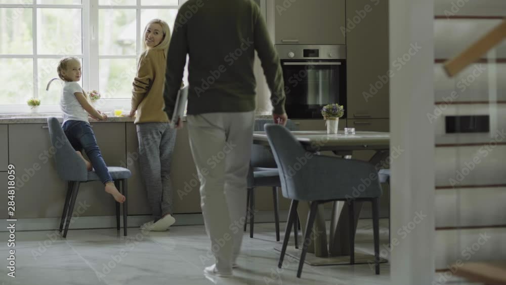 Tracking shot of mother and little daughter cooking together at kitchen counter. Father with laptop walking downstairs to work on computer. Girl giving him glass of orange juice Tracking shot of mother and little daughter cooking together at kitchen counter. Father with laptop walking downstairs to work on computer. Girl giving him glass of orange juice