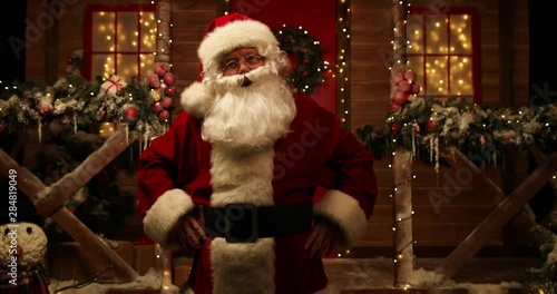 Santa is standing and moving with one hand in front of the porch, decorated for the Christmas. Christmas and New Year time. 