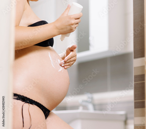 Beautiful pregnant woman in bathroom applying cream on her belly skin.