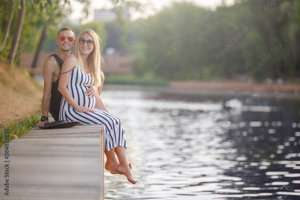 Image of embracing pregnant women and men sitting near river on summer Image of embracing pregnant women and men sitting near river on summer