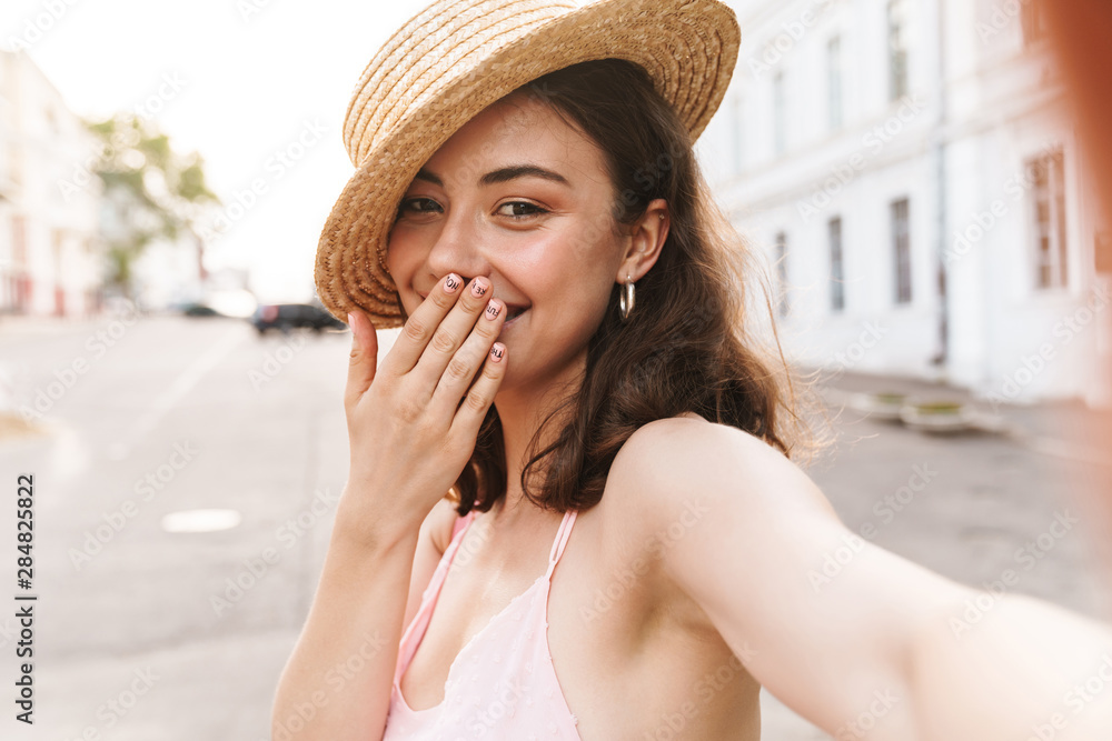 Cheerful young pretty woman in beautiful hat walking by street take selfie by camera.