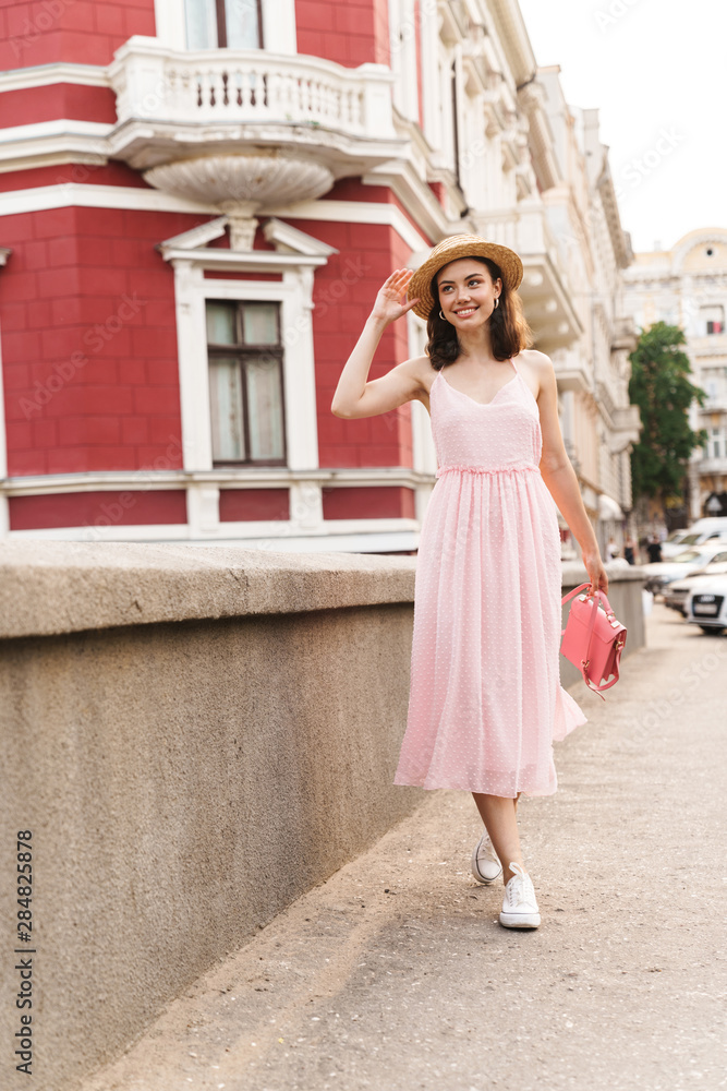 Smiling pleased young pretty woman in beautiful hat walking by street dressed in pink dress.