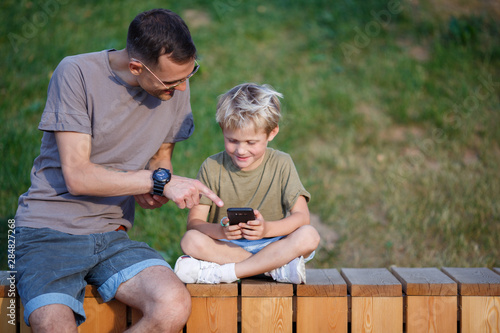 Image of happy man and boy with phones in hands sitting behind wooden fence in park photo