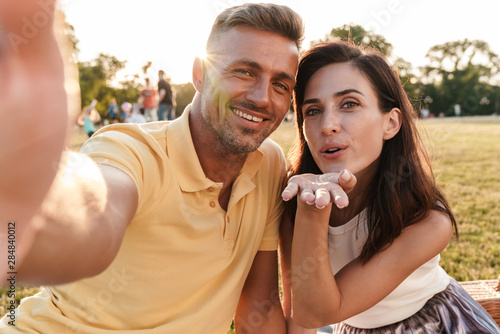 Portrait of pleased middle-aged couple taking selfie photo and sending air kiss while sitting in summer park