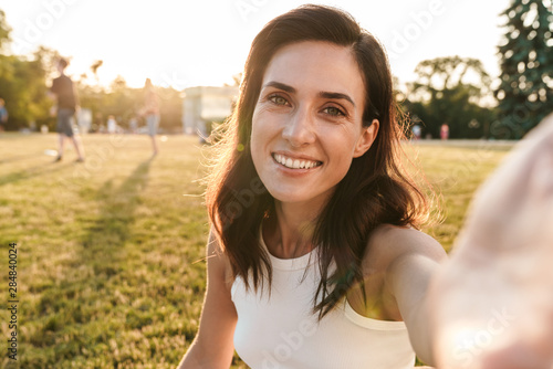 Image of smiling middle-aged woman taking selfie photo while sitting on grass in summer park