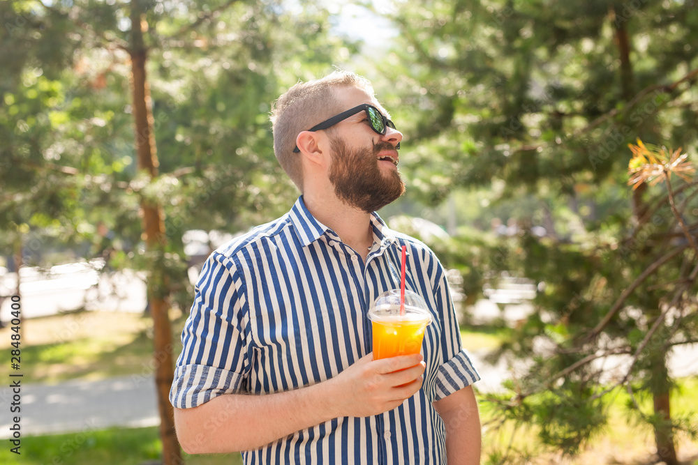 Portrait of a positive cheerful young man with a glass of juice with a straw while walking in the park on a warm sunny summer day. The concept of rest after study and work on weekends. Portrait of a positive cheerful young man with a glass of juice with a straw while walking in the park on a warm sunny summer day. The concept of rest after study and work on weekends.