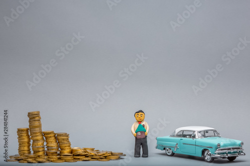 Plasticine man holding a house model in between a big pile of coins and a retro, light blue car figurine, on gray background.