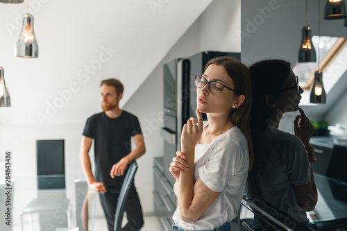 Thoughtful young woman wering wite t-shirt  in glasses with  husband standing behind her background in the kitchen at home and looking at her