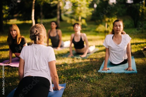 Yoga girls in city park are exercise stretching in pose Urdhva Mukha Shvanasana, Upward Facing Dog. Group of people are meditating on summer sunny morning in park at dawn under guidance of instructor