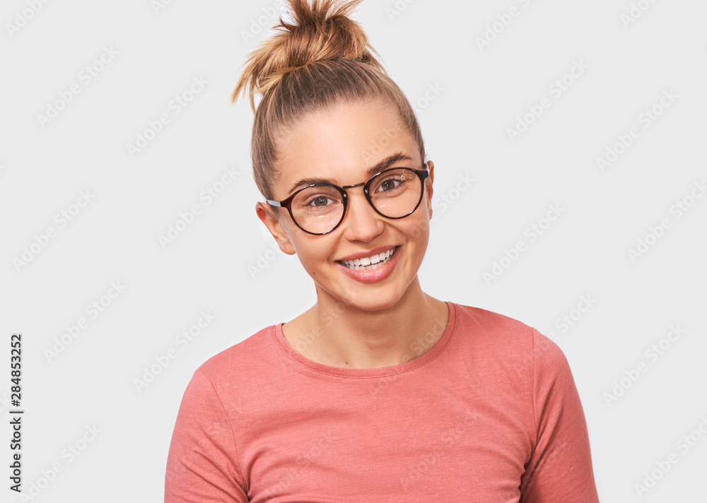 Attractive blonde young woman smiling and wearing round transparent spectacles, casual pink blouse, looks joyful directly into camera, poses against white studio background. People and emotions