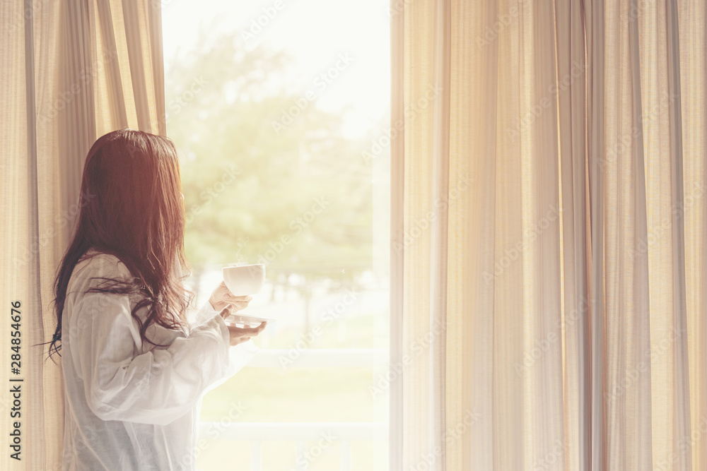 Asian woman in bedroom drinking coffee after wake up and relax mood in holiday near window, sunny morning. Lifestyle Concept.