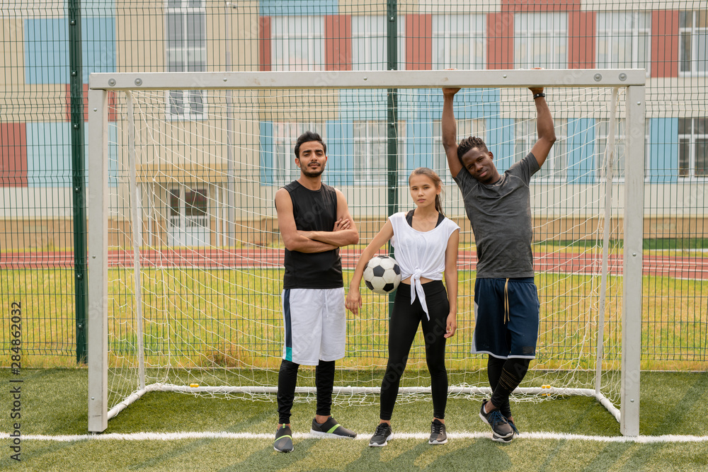 Three young soccer players in sportswear standing by gate on the field