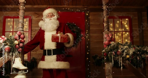 Santa Claus is holding a gift box in his hand and dances on the porch of the house decorated for Christmas. Christmas and New Year time.
