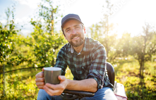 A mature farmer with cup of coffee outdoors in orchard, resting.