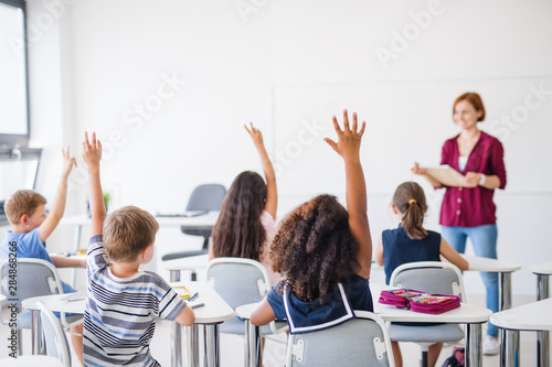 Rear view of school children sitting at the desk in classroom, raising hands.