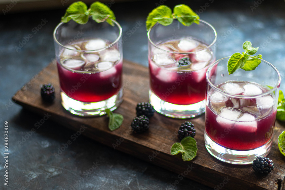 Blackberry tincture in a transparent glass. Ice cubes and pieces of fruit are also in the middle of the dishes. All on a dark background. Slices of lemon and mint leaves serve. Blackberry tincture in a transparent glass. Ice cubes and pieces of fruit are also in the middle of the dishes. All on a dark background. Slices of lemon and mint leaves serve.