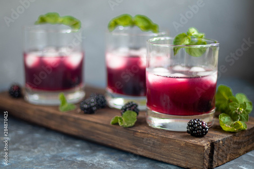 Blackberry tincture in a transparent glass. Ice cubes and pieces of fruit are also in the middle of the dishes. All on a dark background. Slices of lemon and mint leaves serve. 