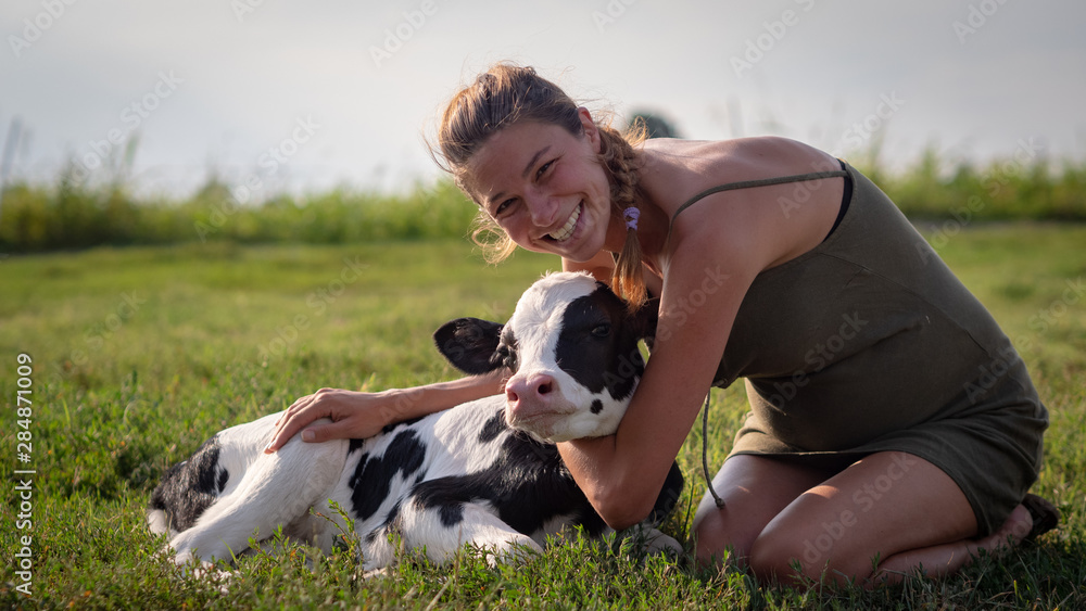 Authentic shot of young woman farmer is caressing an ecologically grown newborn calf used for biological milk products industry on a green lawn of a countryside farm with a sun shining. Authentic shot of young woman farmer is caressing an ecologically grown newborn calf used for biological milk products industry on a green lawn of a countryside farm with a sun shining.