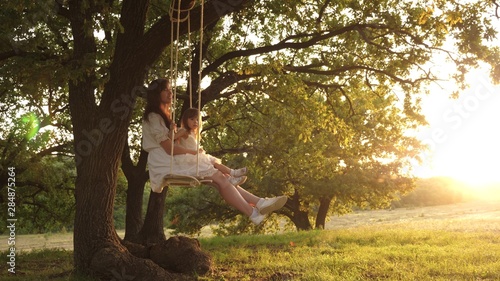 Mom shakes her daughter on swing under tree in sun. mother and baby ride on a rope swing on an oak branch in forest. Girl laughs, rejoices. Family fun in park, in nature. warm summer day.