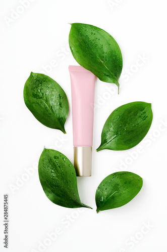 Cosmetology tube with green leaves on white background. Top class facial care serum with decorative fresh ficus foliage and dew drops botanical backdrop. Eye cream, concealer in pink tube promo