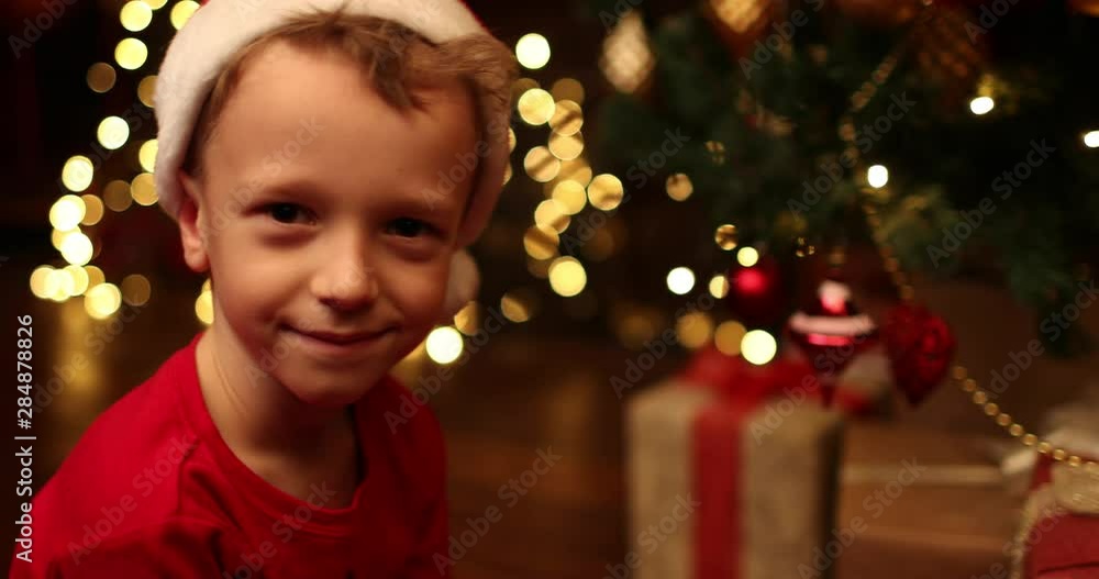 A little boy in a Christmas cap is sitting near decorated Christmas tree. Merry Christmas, happy New Year. Luxurious apartment with christmas lights and decorations. Miracle time. A little boy in a Christmas cap is sitting near decorated Christmas tree. Merry Christmas, happy New Year. Luxurious apartment with christmas lights and decorations. Miracle time.