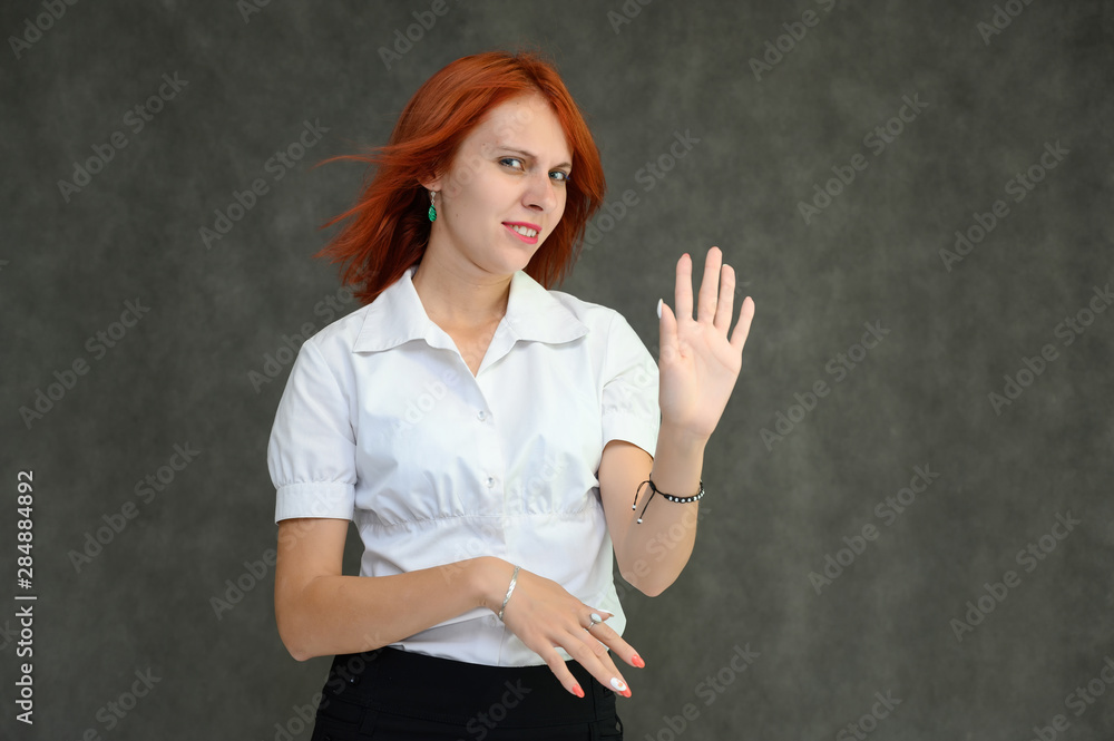 Photo Portrait of a cute girl woman with bright red hair manager in a white shirt on a gray background in the studio. He talks, shows his hands in front of the camera with emotions.